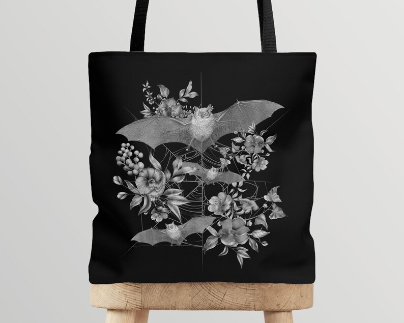 May include: Black tote bag with a white design of bats and flowers. The bats are flying in a web with flowers and leaves around them.