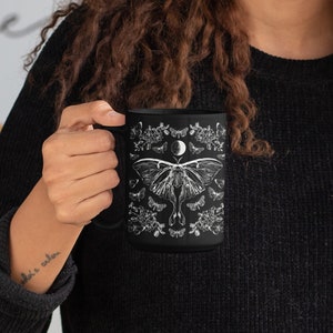 May include: Black ceramic mug featuring a detailed white illustration of a moth, butterflies, flowers, and a crescent moon. The mug has a large handle and is held by a person wearing a black sweater. The design is symmetrical and nature-themed.