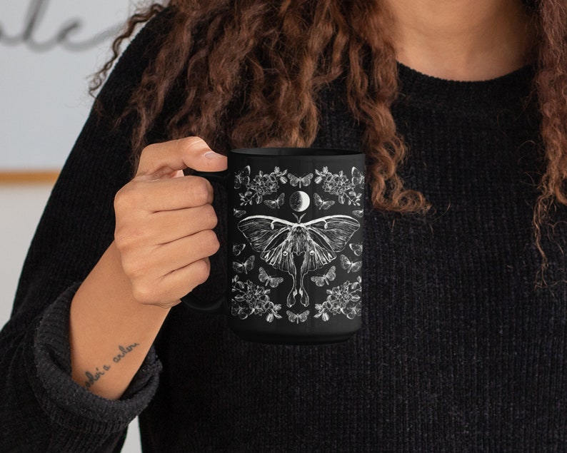 May include: Black ceramic mug featuring a detailed white illustration of a moth, butterflies, flowers, and a crescent moon. The mug has a large handle and is held by a person wearing a black sweater. The design is symmetrical and nature-themed.