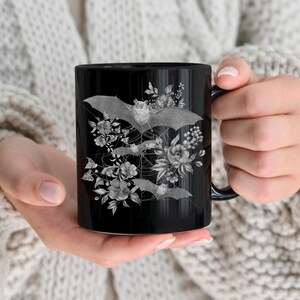 May include: Black ceramic mug with a white design of bats and flowers. The bats are flying in a circular pattern around the flowers.