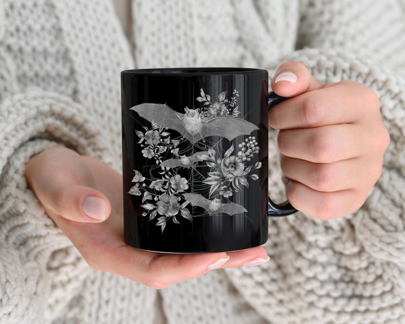 May include: Black ceramic mug with a white design of bats and flowers. The bats are flying in a circular pattern around the flowers.