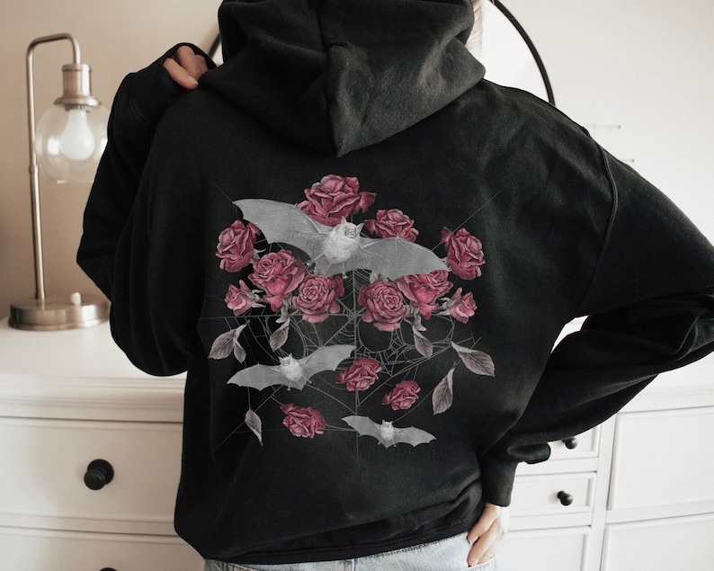May include: Black hoodie featuring a floral and bat design. The graphic includes pink roses, gray bats, and spiderwebs. The design is centered on the back of the hoodie. The hoodie has a drawstring hood and long sleeves.