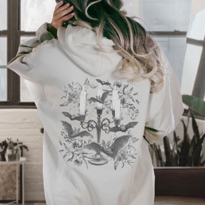 May include: A white hoodie featuring a black and white graphic print of a candelabra with bats and flowers. The design is centred on the back of the hoodie.