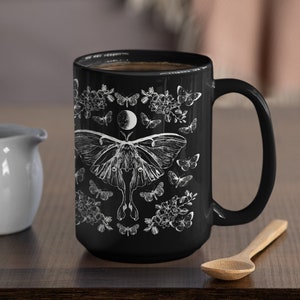 May include: Black ceramic mug with a white illustration of a moth, butterflies, flowers, and a crescent moon. The mug is filled with dark liquid and has a large handle. A small white creamer and wooden spoon are next to the mug.