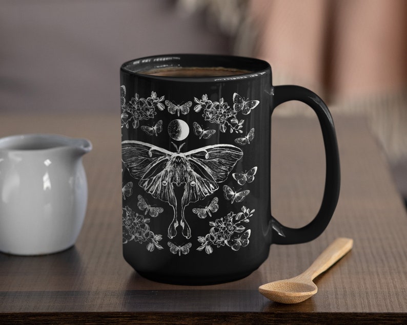 May include: Black ceramic mug with a white illustration of a moth, butterflies, flowers, and a crescent moon. The mug is filled with dark liquid and has a large handle. A small white creamer and wooden spoon are next to the mug.