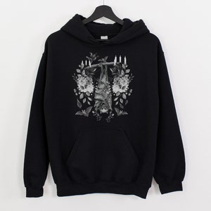 May include: Black hoodie featuring a grayscale illustration of a bat hanging upside down, surrounded by flowers and candles. The design is centered on the front of the hooded sweatshirt, which has a kangaroo pocket.