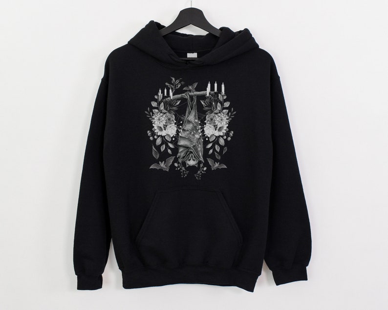 May include: Black hoodie featuring a grayscale illustration of a bat hanging upside down, surrounded by flowers and candles. The design is centered on the front of the hooded sweatshirt, which has a kangaroo pocket.