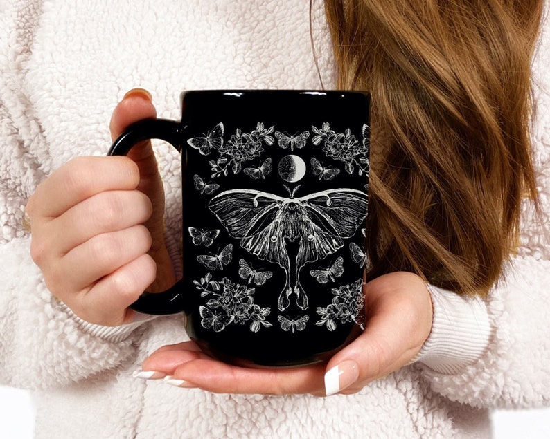 May include: Black ceramic mug featuring a white illustration of a moth, butterflies, flowers, and a crescent moon. The mug has a black handle and a glossy finish. The design is symmetrical and detailed, creating a gothic or celestial theme.