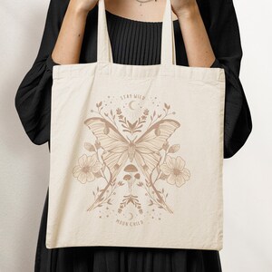 May include: A beige tote bag featuring a brown illustration of a moth, flowers, and mushrooms. The text "Stay Wild Moon Child" is printed on the bag.