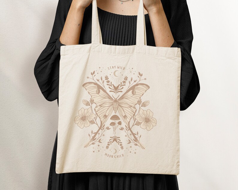 May include: A beige tote bag featuring a brown illustration of a moth, flowers, and mushrooms. The text "Stay Wild Moon Child" is printed on the bag.