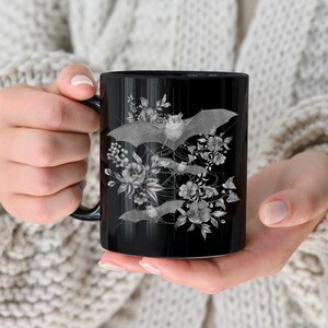 May include: Black ceramic mug with a white design of bats and flowers. The bats are flying in a circular pattern around the flowers.