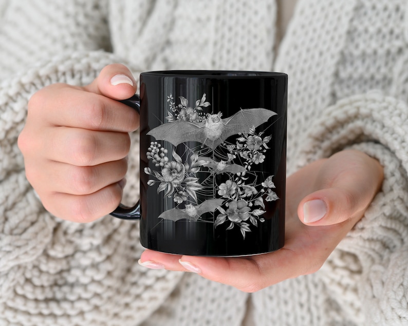 May include: Black ceramic mug with a white design of bats and flowers. The bats are flying in a circular pattern around the flowers.