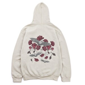 May include: A beige hoodie featuring a graphic design on the back. The design includes gray bats, dark red roses, and a spiderweb-like pattern. The hoodie has a hood and a relaxed fit, suitable for casual wear.