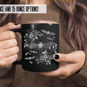 Pu&ograve; includere: Tazza in ceramica nera con un design bianco di pipistrelli che volano intorno a una ragnatela con rose. Il testo "11 OUNCE AND 15 OUNCE OPTIONS!" &egrave; in cima alla tazza.