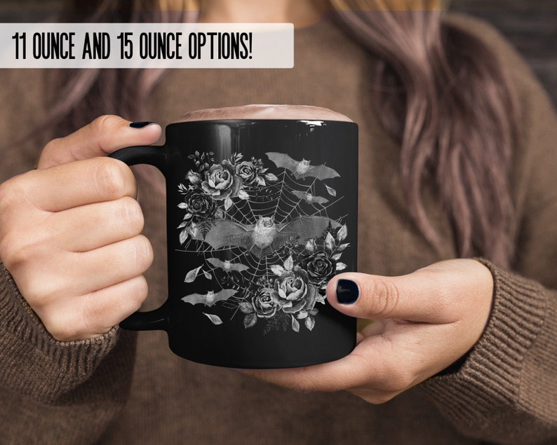 Pu&ograve; includere: Tazza in ceramica nera con un design bianco di pipistrelli che volano intorno a una ragnatela con rose. Il testo "11 OUNCE AND 15 OUNCE OPTIONS!" &egrave; in cima alla tazza.