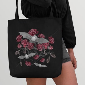 May include: Black tote bag with a design of bats and red roses on a black background. The bats are flying around the roses, which are arranged in a cluster. The design is surrounded by a web-like pattern.