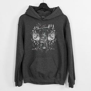May include: Dark gray hoodie with a floral and bat design. The graphic features a bat hanging upside down from a branch, surrounded by flowers and candles. The hoodie has a front pocket and a hood.