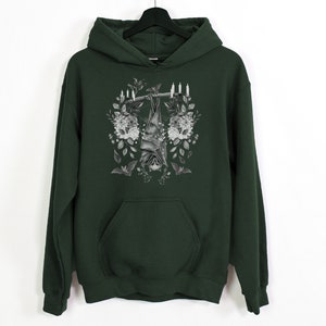 May include: Dark green hoodie with a black and white bat graphic hanging from a branch, surrounded by flowers and candles. The hoodie has a front pocket and a hood, and is hanging on a black hanger.