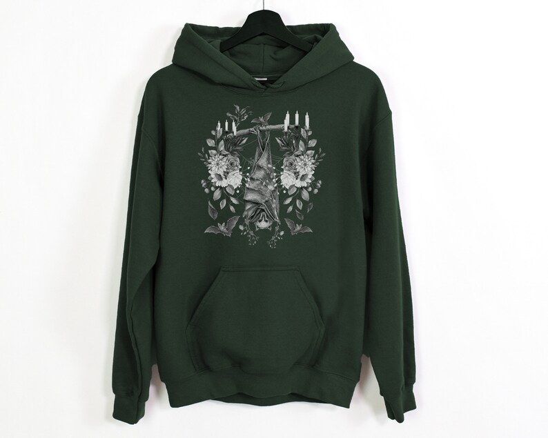 May include: Dark green hoodie with a black and white bat graphic hanging from a branch, surrounded by flowers and candles. The hoodie has a front pocket and a hood, and is hanging on a black hanger.