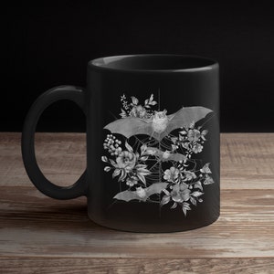 May include: Black ceramic mug with a white design of bats and flowers. The bats are flying in a web-like pattern with flowers and leaves surrounding them.