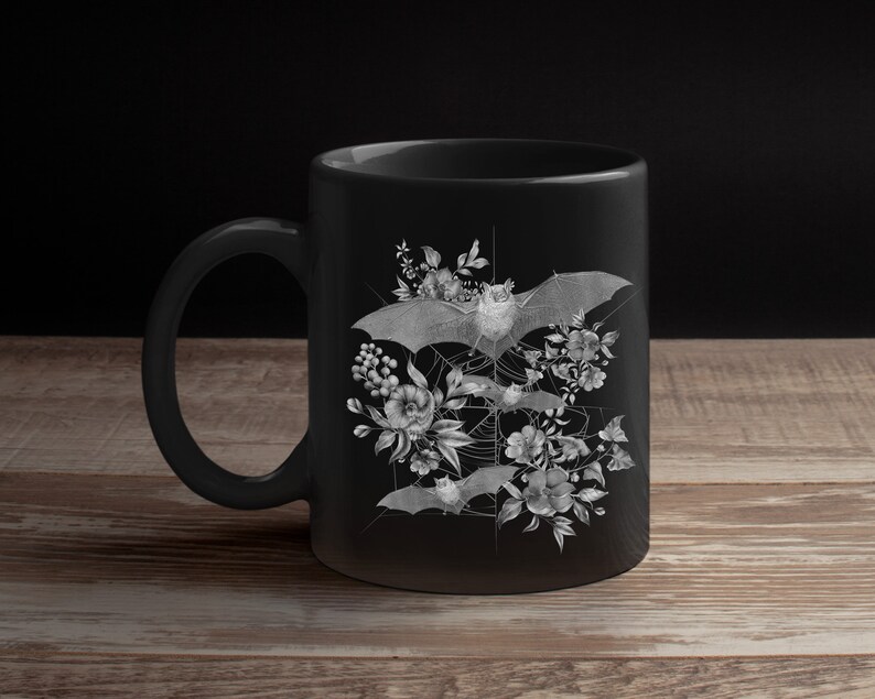 May include: Black ceramic mug with a white design of bats and flowers. The bats are flying in a web-like pattern with flowers and leaves surrounding them.