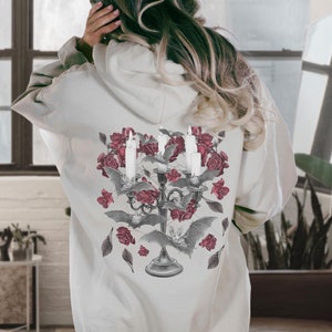 May include: A white hoodie with a graphic print of a candelabra with candles, roses, and bats. The graphic is in shades of gray and red.