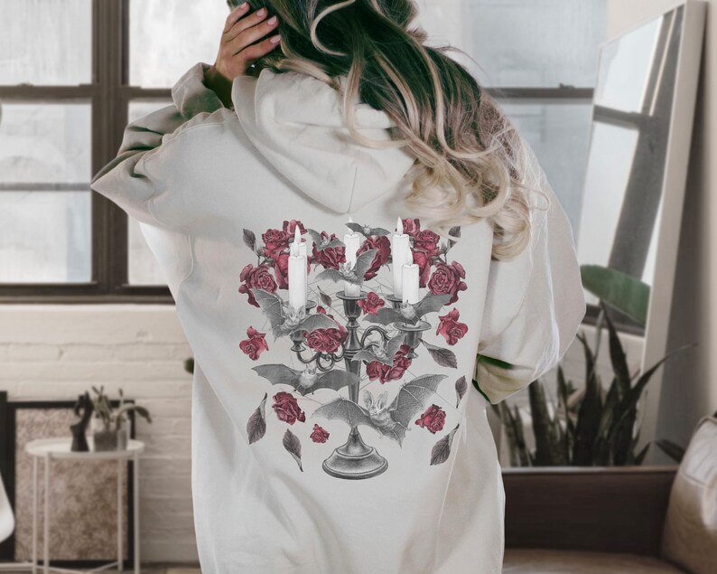 May include: A white hoodie with a graphic print of a candelabra with candles, roses, and bats. The graphic is in shades of gray and red.