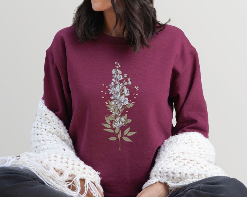 May include: A maroon sweatshirt with a white floral design on the front. The design features a cluster of white flowers with green leaves.