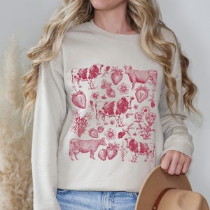 May include: A cream-colored sweatshirt with a red graphic print of cows and strawberries. The graphic is a vintage-style illustration.