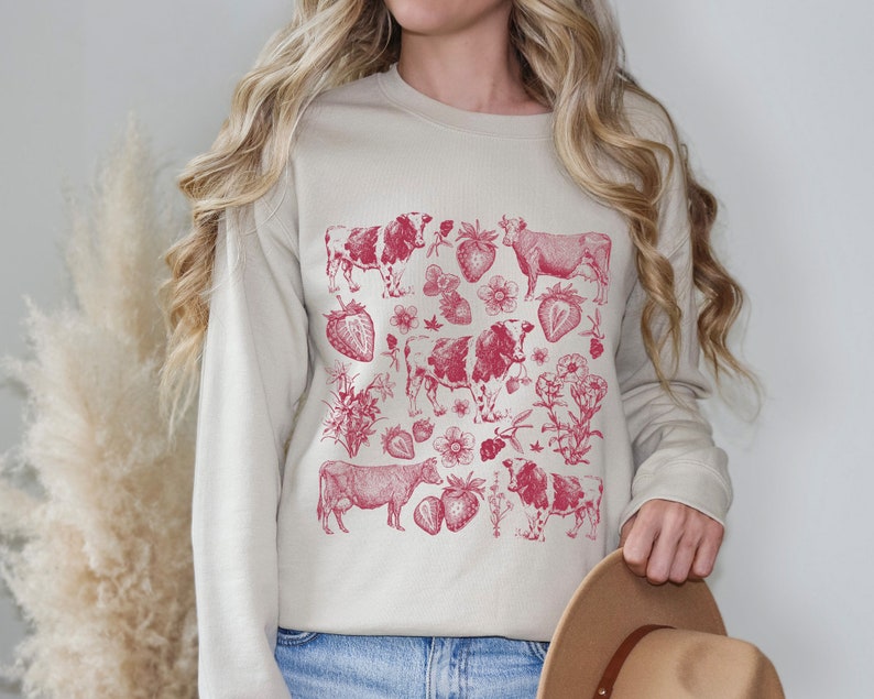 May include: A cream-colored sweatshirt with a red graphic print of cows and strawberries. The graphic is a vintage-style illustration.