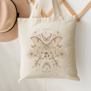 May include: A beige canvas tote bag featuring a brown butterfly and floral design. The bag has the text "Stay Wild Moon Child" printed on it.