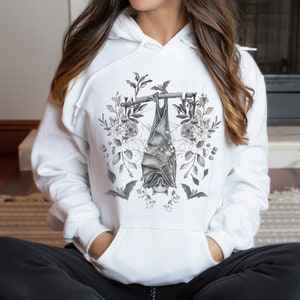 May include: White hoodie featuring a grayscale bat design. The bat hangs upside down from a branch, surrounded by floral and botanical elements. The design includes bats, flowers, and leaves, creating a gothic and nature-inspired aesthetic.