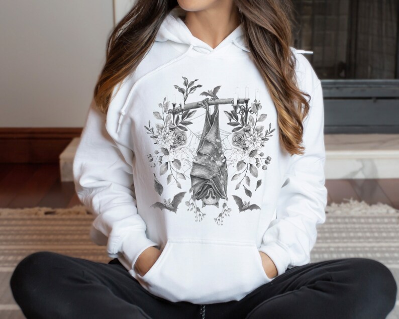 May include: White hoodie featuring a grayscale bat design. The bat hangs upside down from a branch, surrounded by floral and botanical elements. The design includes bats, flowers, and leaves, creating a gothic and nature-inspired aesthetic.