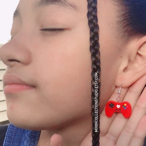 May include: Red video game controller earrings with silver hooks. The controller has a black cross, a white heart, and colorful buttons. The earrings are shown next to a person's face. Text on the image says MOONCOLLECTIONSTUDIO.ETSY.COM.