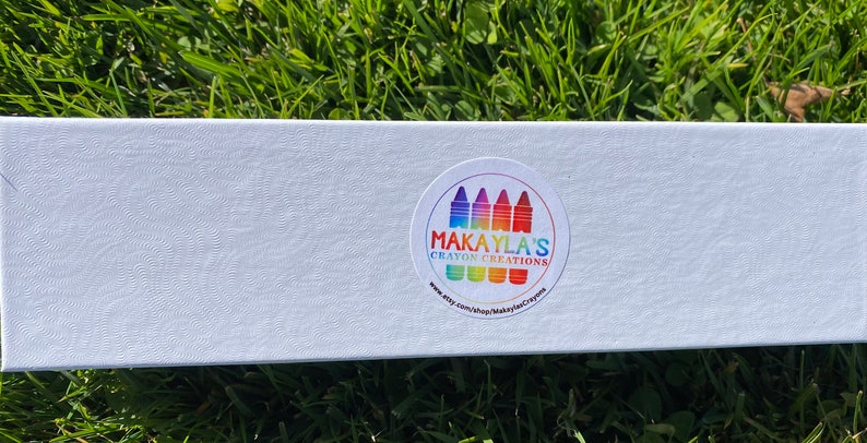 K&ouml;nnte beinhalten: Wei&szlig;e Box mit einem bunten Aufkleber, auf dem "Makayla's Crayon Creations" und eine Website-Adresse stehen. Der Aufkleber hat ein Regenbogen-Design mit Buntstiften.