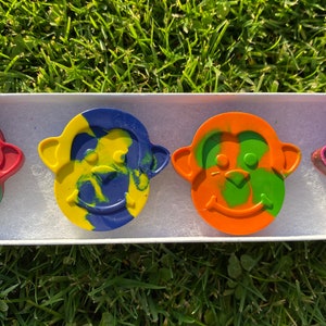 May include: A set of four monkey-shaped crayons in a white box, each with a unique color combination. The crayons are green and red, yellow and blue, orange and green, and brown and pink.