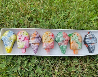 May include: A set of eight colorful ice cream cone shaped crayons in a white box. The crayons are decorated with glitter and have a variety of colors, including blue, yellow, pink, purple, orange, green, and black.