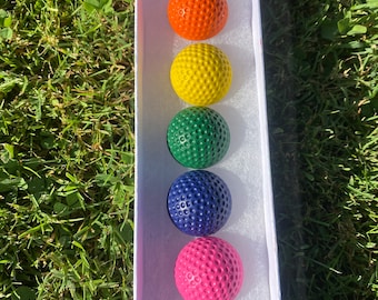 May include: A set of seven golf balls in a white box. The balls are arranged in a row from top to bottom, with the colors red, orange, yellow, green, blue, pink, and purple.