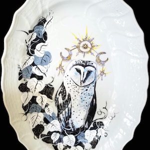 May include: A white porcelain oval platter featuring a hand-painted design of a barn owl with a sun and moon above it. The owl is surrounded by black and white vines.