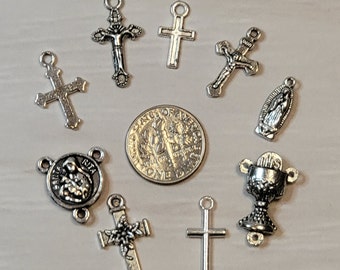 May include: A collection of silver-toned religious charms, including crosses, a chalice, and a depiction of the Virgin Mary. The charms are arranged on a white surface with a US dime for scale.