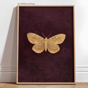 May include: A framed print featuring a gold-colored butterfly against a deep burgundy textured background. The butterfly is centered, with detailed wing patterns. The frame is a light wood color, and the print is leaning against a white wall.