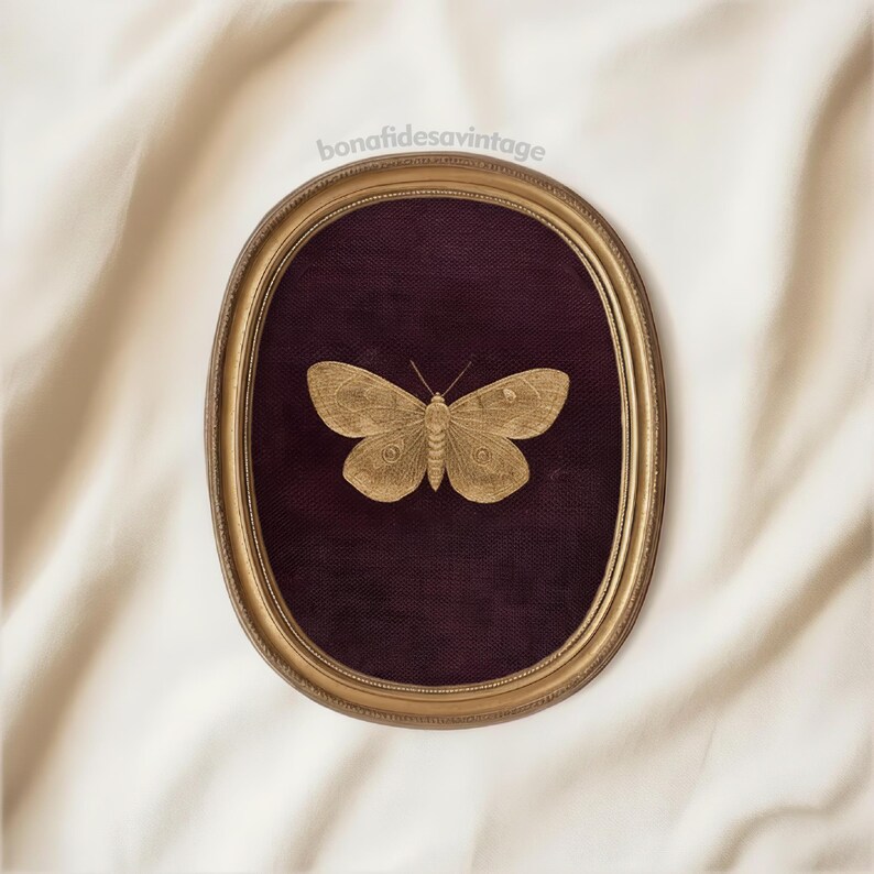 May include: A gold-colored moth framed in an oval, gold-toned frame. The moth is centered on a dark purple background. The frame is set against a white, textured fabric. The text "bonafidesvintage" is in the upper left corner.
