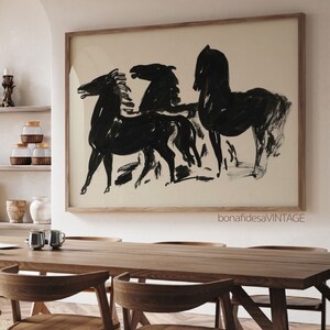 May include: Black and white abstract painting of three horses in a minimalist style. The painting is framed in a natural wood frame and hangs on a white wall above a wooden table.