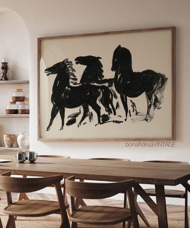 May include: Black and white abstract painting of three horses in a minimalist style. The painting is framed in a natural wood frame and hangs on a white wall above a wooden table.