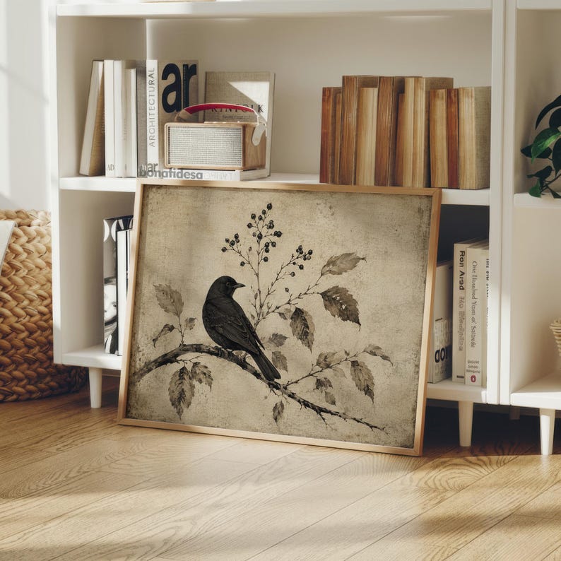 Peut inclure: Une impression d'art encadr&eacute;e carr&eacute;e repr&eacute;sentant un oiseau noir perch&eacute; sur une branche avec des feuilles et des baies sur un fond beige textur&eacute;. L'&oelig;uvre pr&eacute;sente une palette de couleurs sourdes et un aspect vieilli, dans un cadre en bois clair.