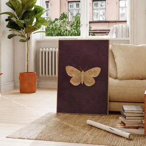 May include: A framed artwork featuring a gold-colored butterfly against a deep burgundy background. The butterfly is centered, with detailed wing patterns. The artwork is displayed in a living room setting with a neutral-toned sofa and a potted plant.