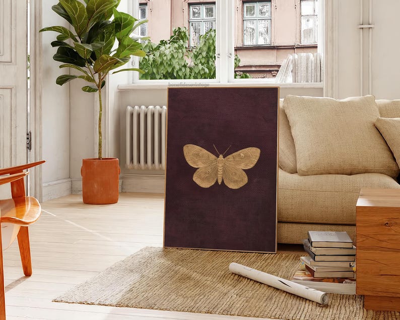 May include: A framed artwork featuring a gold-colored butterfly against a deep burgundy background. The butterfly is centered, with detailed wing patterns. The artwork is displayed in a living room setting with a neutral-toned sofa and a potted plant.