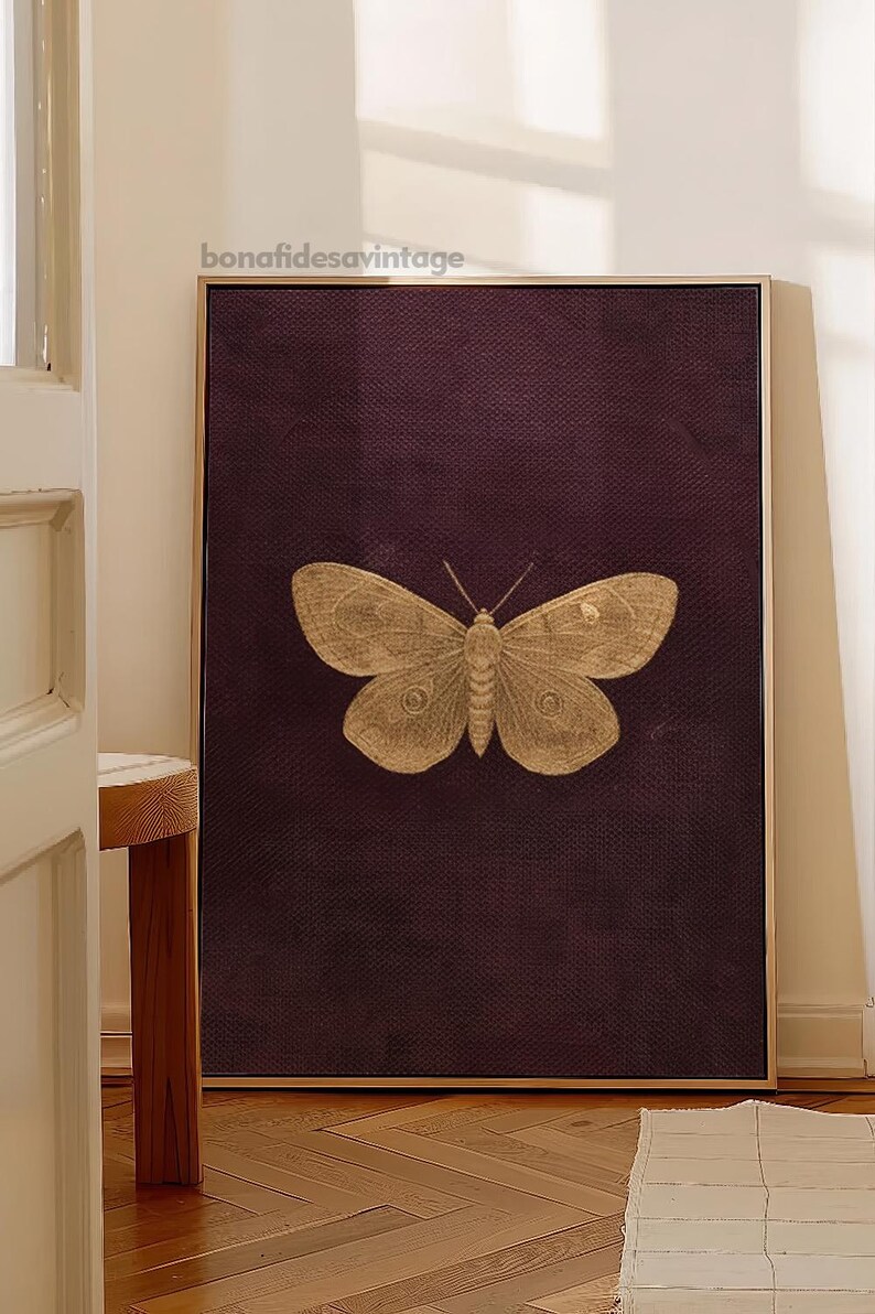May include: A framed print featuring a detailed illustration of a butterfly in shades of gold. The butterfly is centered on a dark burgundy textured background. The frame is a light wood color, and the print is displayed in a room with natural light.
