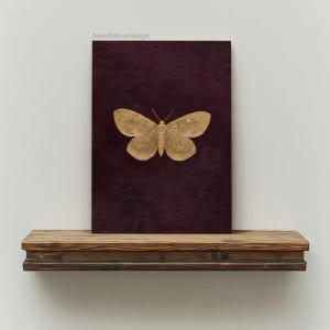 May include: A gold-colored butterfly is centered on a dark burgundy canvas. The canvas is displayed on a rustic, dark wood shelf. The image has a vintage aesthetic. The text "bonafidesavintage" is in the upper left corner.