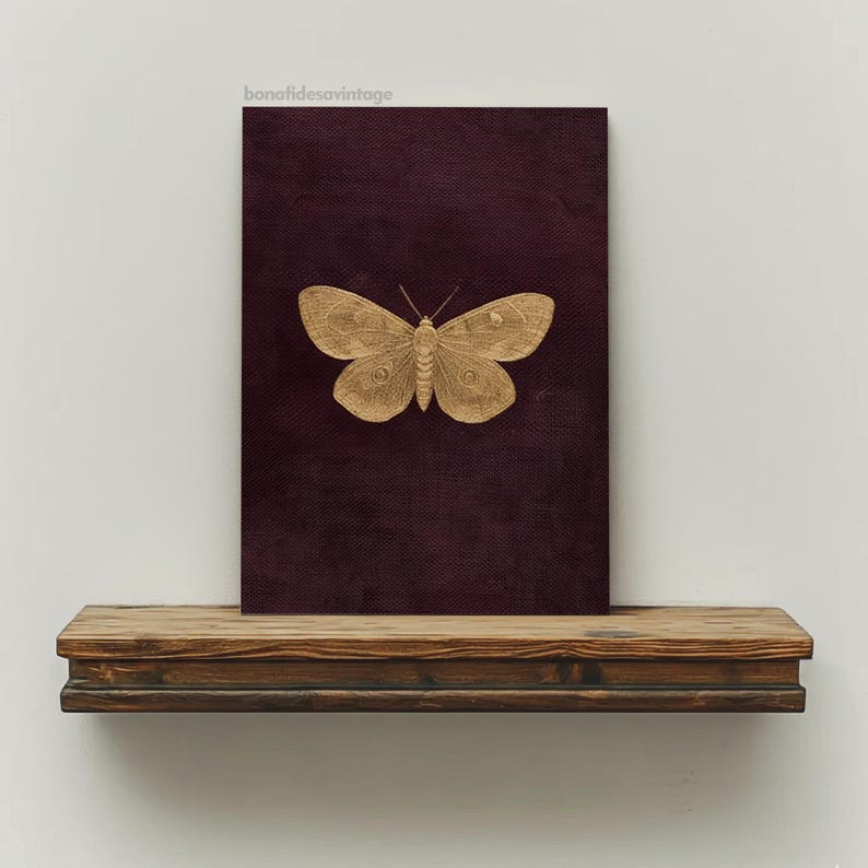 May include: A gold-colored butterfly is centered on a dark burgundy canvas. The canvas is displayed on a rustic, dark wood shelf. The image has a vintage aesthetic. The text "bonafidesavintage" is in the upper left corner.